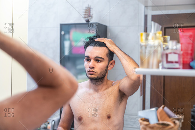 Young shirtless unshaven guy looking in the mirror  and examining hair during daily morning routine at home