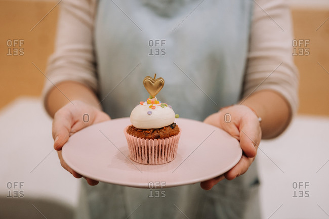 Crop unrecognizable female in apron holding plate with yummy sweet cupcake decorated with burning candle