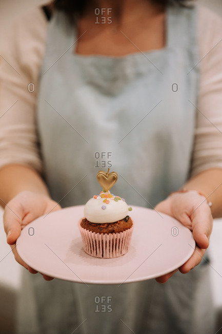 Crop unrecognizable female in apron holding plate with yummy sweet cupcake decorated with burning candle