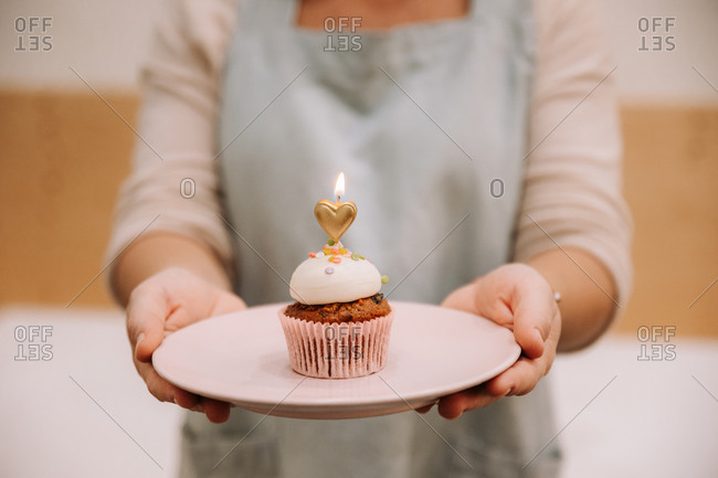 Crop unrecognizable female in apron holding plate with yummy sweet cupcake decorated with burning heart shaped candle