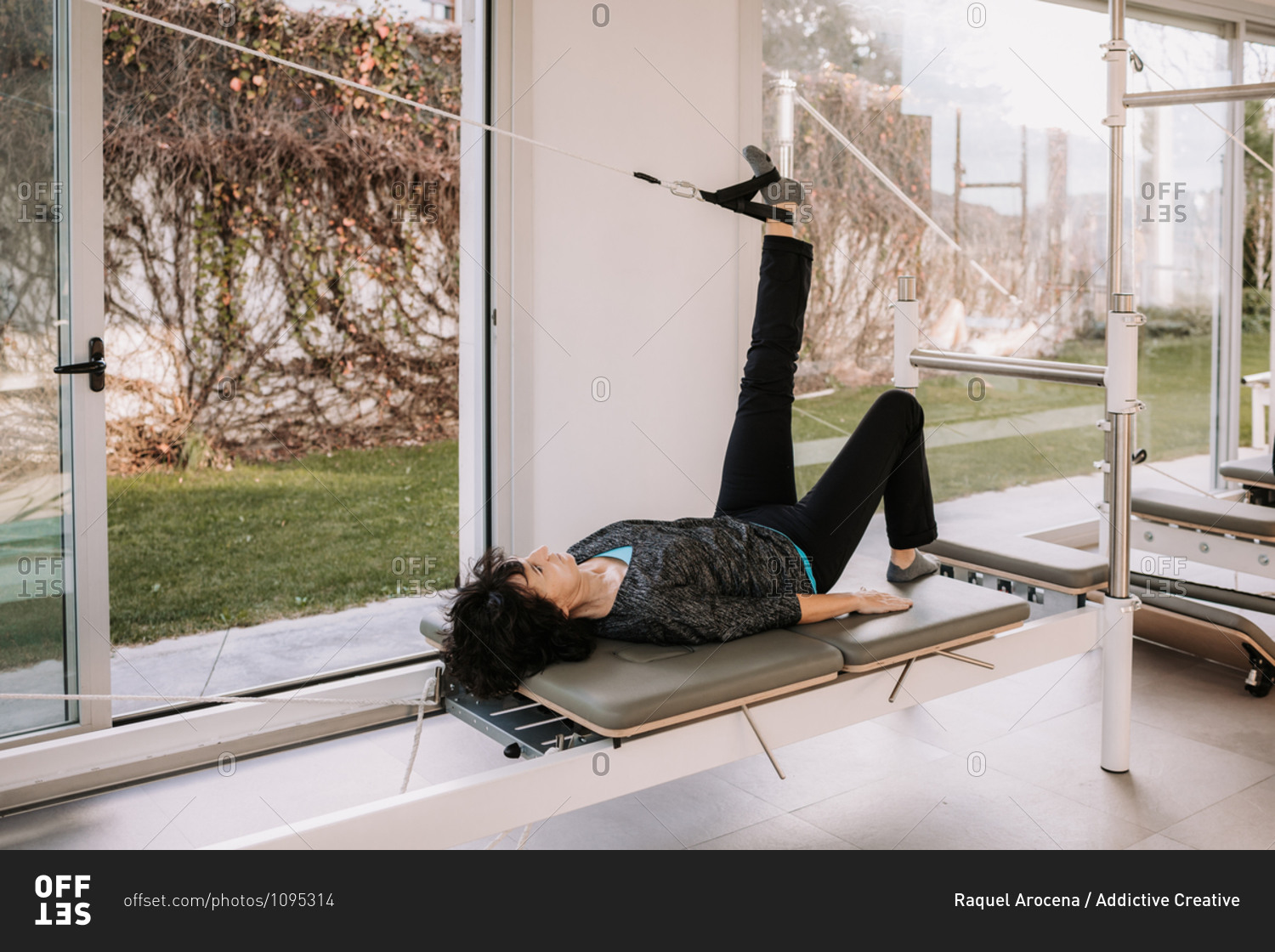 Side view of company of people in activewear lying on pilates reformer with straps in Bridge pose with raised leg and doing yoga in gym together while stretching body