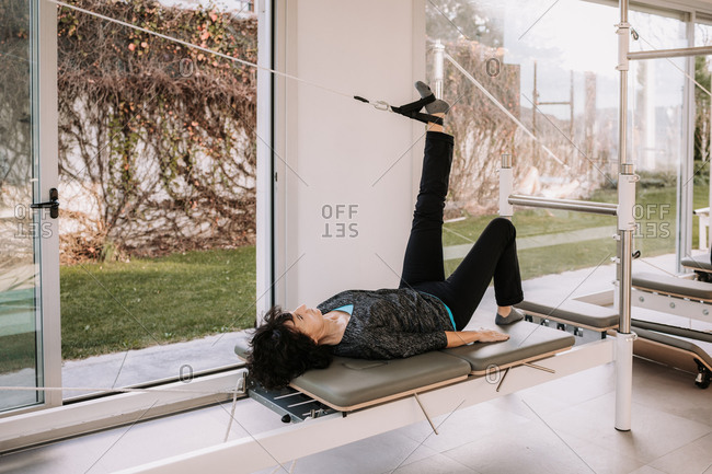 Side view of company of people in activewear lying on pilates reformer with straps in Bridge pose with raised leg and doing yoga in gym together while stretching body