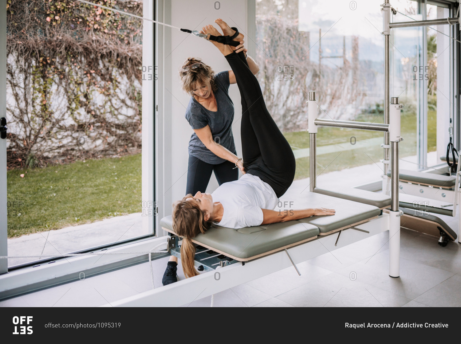 Female in sportswear lying on pilates cadillac and stretching legs with straps under control of professional trainer in modern gym