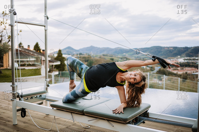 Cheerful female in sportswear practicing yoga in Side Plank on the Knee pose and stretching arm with strap while doing pilates exercises on reformer on terrace
