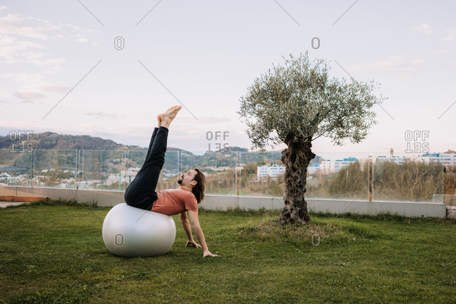 Flexible male in activewear leaning on fit balls and doing yoga in Camatkarasana on green lawn during pilates training