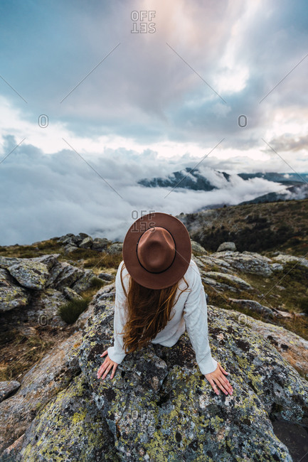 Back view of faceless female tourist in hat sitting on stone and observing scenic view of mountains on cloudy day