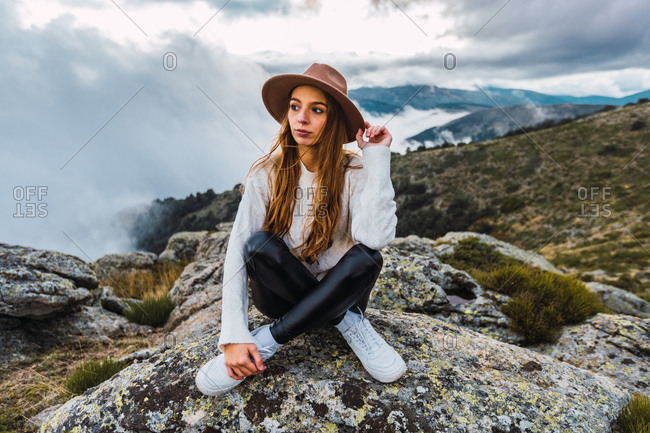Female tourist in hat sitting on stone and observing scenic view of mountains on cloudy day looking away