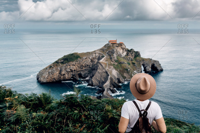 Back view of unrecognizable male tourist standing from above observing scenery of Gaztelugatxe island in sea during summer vacation