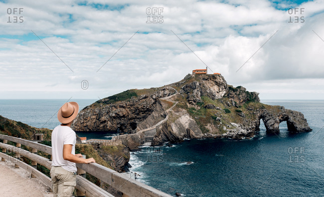 Back view of unrecognizable male tourist standing on embankment and observing scenery of Gaztelugatxe island in sea during summer vacation
