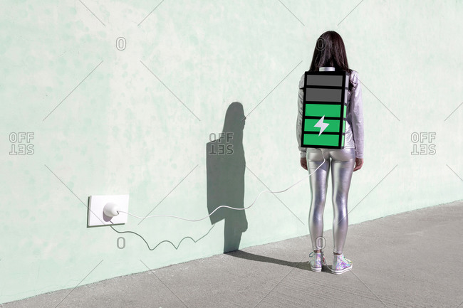 Back view of unrecognizable female with long dark hair in stylish outfit standing on street near wall with battery shaped backpack connected to charger