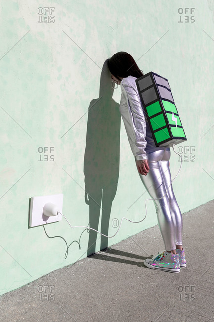 Side view of unrecognizable exhausted female in futuristic outfit leaning head on wall while standing on street and recharging energy