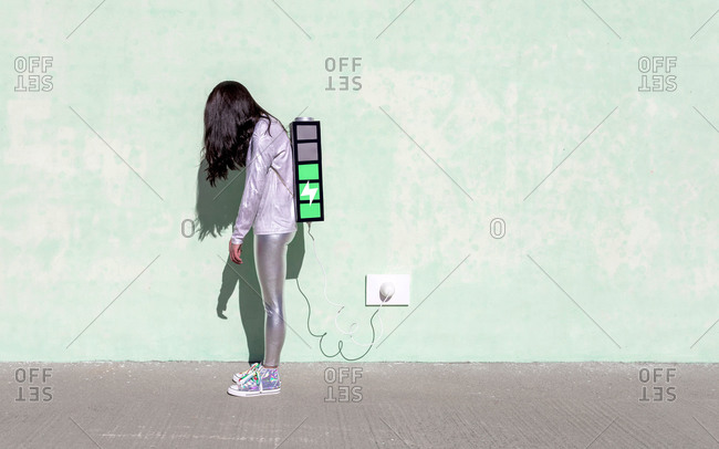 Side view of young fit female in trendy outfit standing on street with creative backpack connected to charger and having phone conversation