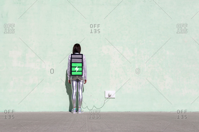 Back view of unrecognizable female with long dark hair in stylish outfit standing on street near wall with battery shaped backpack connected to charger