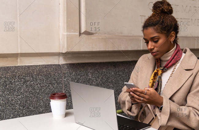 Stylish young African American female freelancer in trendy outfit talking on mobile phone and working remotely on laptop in cafe