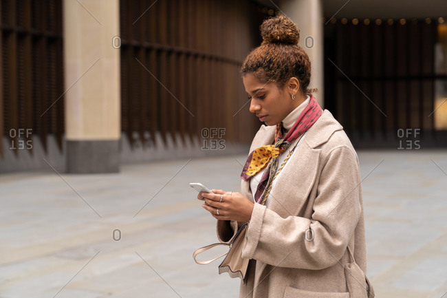 Side view of young African American female entrepreneur with curly hair in elegant coat using smartphone while crossing road in London with laptop in hand