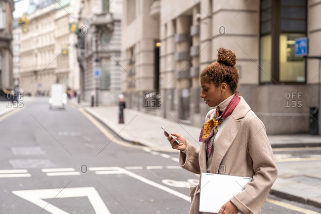 Side view of young African American female entrepreneur with curly hair in elegant coat using smartphone while crossing road in London with laptop in hand