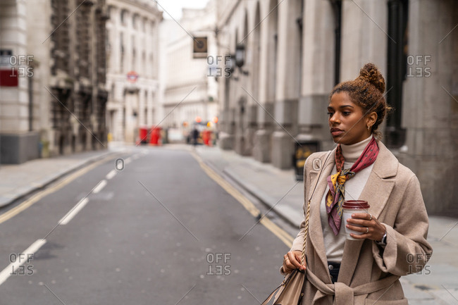Confident young African American female entrepreneur in fashionable outfit listening to music with wireless earphones and drinking takeaway coffee while walking on city street during break
