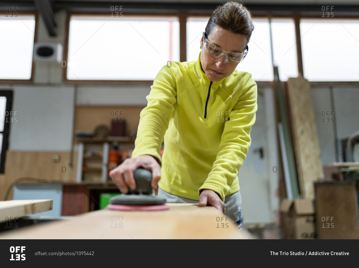 Low angle of professional mature female master in casual clothes and goggles polishing wooden plank using random orbital sander in joinery