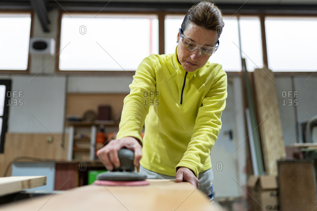 Low angle of professional mature female master in casual clothes and goggles polishing wooden plank using random orbital sander in joinery