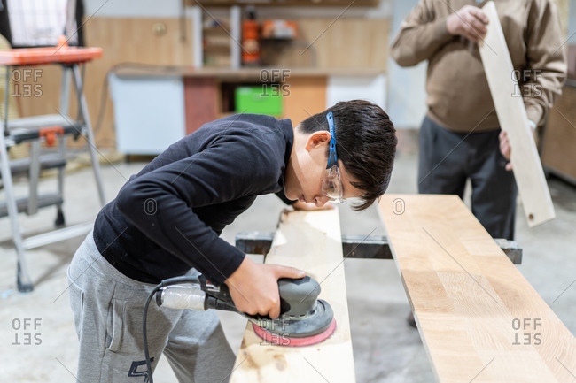 Side view of concentrated boy in casual clothes and goggles smoothing wooden panel with random orbital sander while helping crop anonymous craftsman at workshop