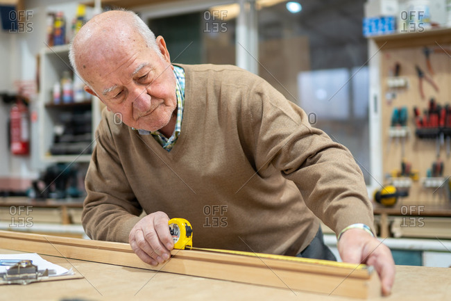 Elderly male woodworker measuring material with tape placed on machine in joiner