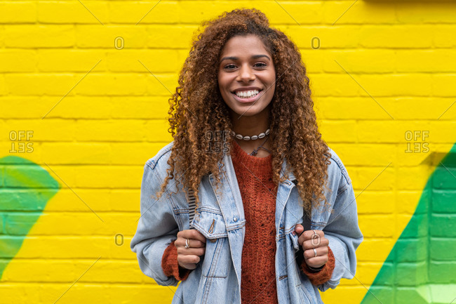 Optimistic young black female millennial with curly hair in stylish clothes smiling while looking at camera against yellow wall