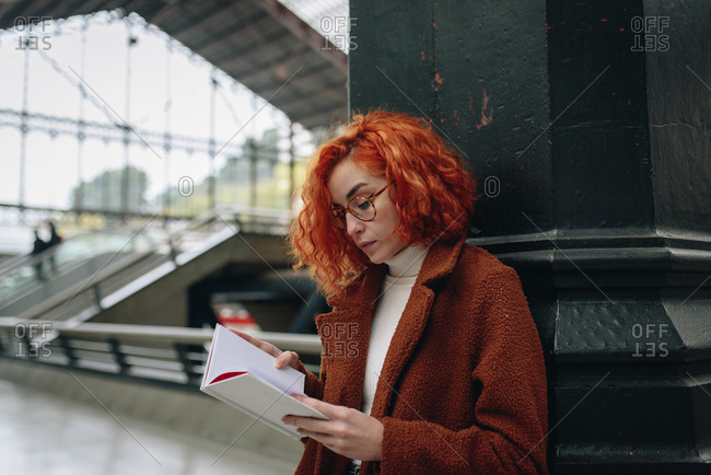 Tranquil female with red hair standing at railroad station and reading interesting book