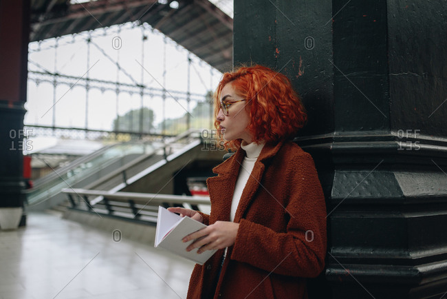Tranquil female with red hair standing at railroad station and reading interesting book