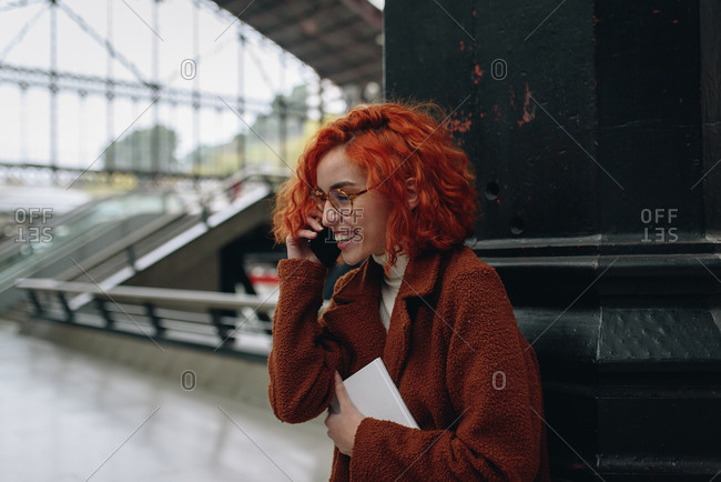 Delighted female with red hair and in autumn coat standing at railroad station and speaking on mobile phone while enjoying conversation