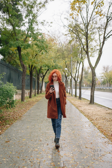 Optimistic female with red hair and in autumn coat standing on alley in park and enjoying smartphone conversation with friend