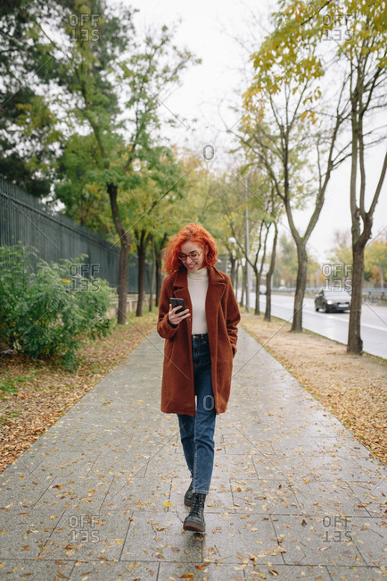 Optimistic female with red hair and in autumn coat standing on alley in park and enjoying smartphone conversation with friend