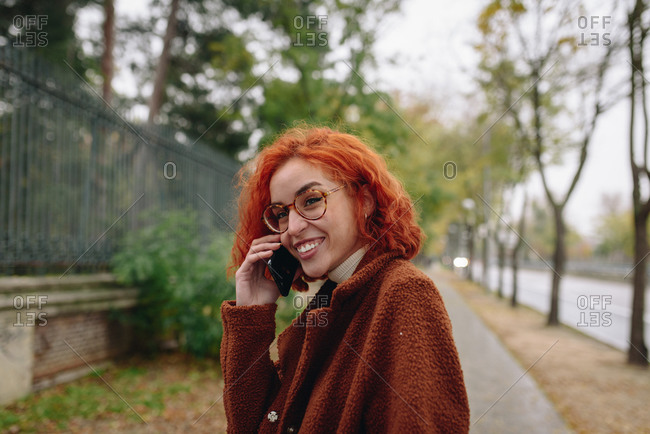 Delighted female with red hair and in autumn coat standing at the park and speaking on mobile phone while enjoying conversation