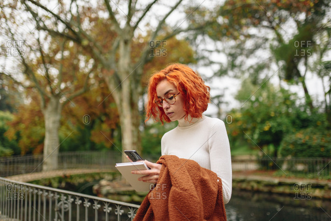 Tranquil female with red hair standing in park and surfing Internet on smartphone while enjoying weekend