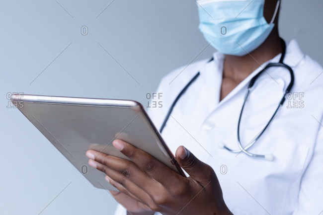 Mixed race female doctor wearing face mask standing and using digital tablet. medical professional healthcare worker hygiene during coronavirus covid 19 pandemic.