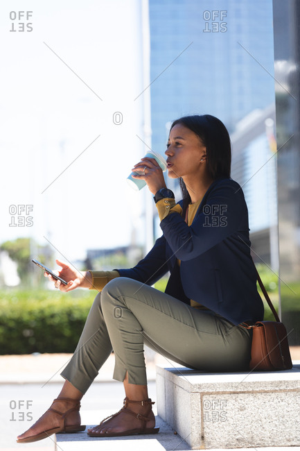 African American woman with smartphone drinking coffee while sitting at corporate park. lifestyle living during coronavirus covid 19 pandemic.
