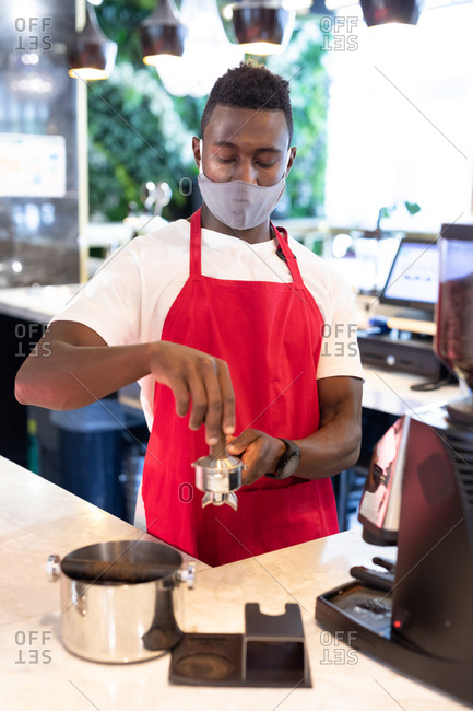 Portrait of African American male barista wearing face mask using coffee machine looking at camera. health and hygiene in business during coronavirus covid 19 pandemic.