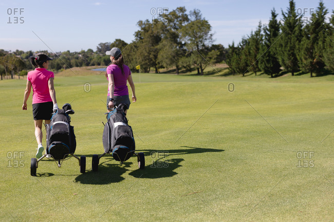 Two caucasian women walking across golf course talking pulling golf bags on wheels. sport leisure hobbies golf healthy outdoor lifestyle.