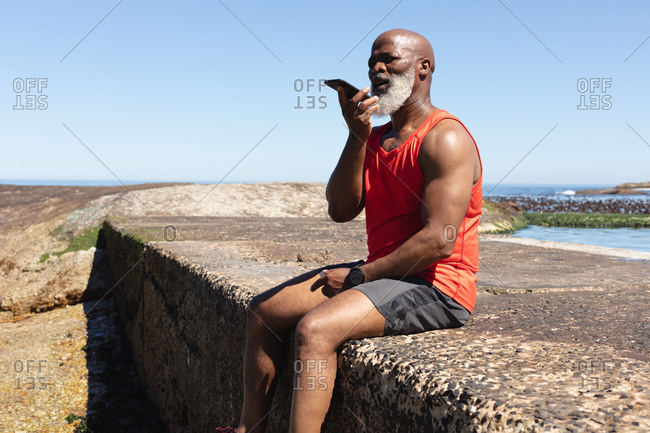 Fit senior African American man sitting on wall by the sea talking on smartphone. healthy retirement outdoor fitness lifestyle.