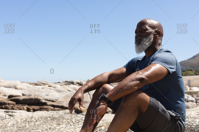 Fit senior African American man sitting on rocky coast looking away. healthy retirement outdoor fitness lifestyle.