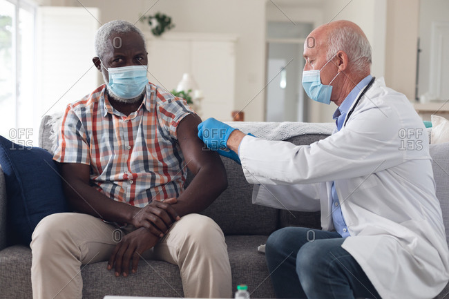 Senior caucasian male doctor vaccinating male patient both wearing face masks at home. healthcare hygiene protection during coronavirus covid 19 pandemic.