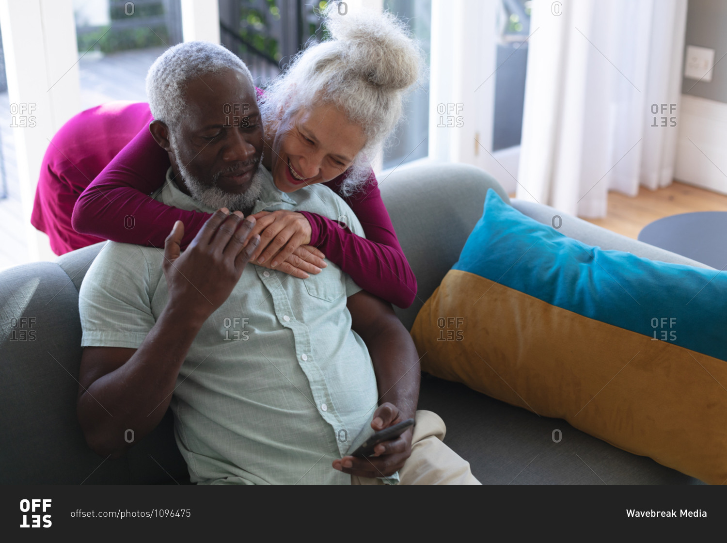 Senior mixed race couple embracing looking at smartphone together in living room. staying at home in self isolation during quarantine lockdown.