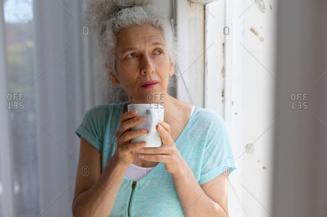 Senior caucasian woman standing by window drinking cup of coffee at home. staying at home in self isolation during quarantine lockdown.
