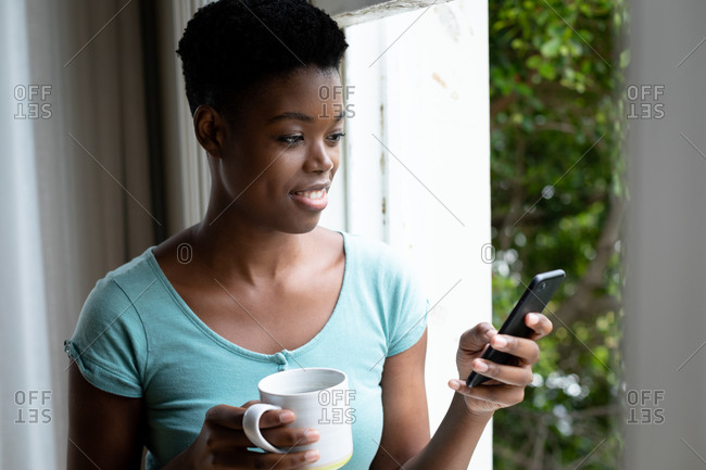 Portrait of African American woman holding coffee cup using smartphone at home. staying at home in self isolation in quarantine lockdown