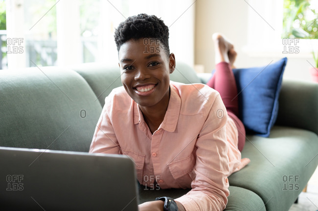 Portrait of African American woman lying on couch using laptop working from home. looking at camera and smiling. staying at home in self isolation during quarantine lockdown.