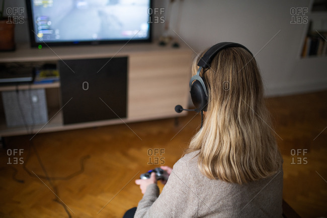 Rear view of a blonde woman sitting on chair in living room wearing a headset and playing video games