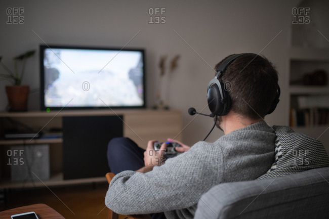 Rear view of a man sitting on chair in living room wearing a headset and playing video games