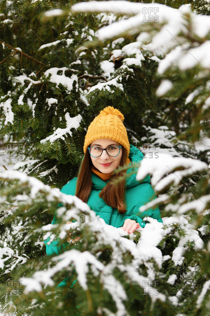 The young female woman in a green winter jacket is standing and touching the snowy trees in the park or forest that is full of snow