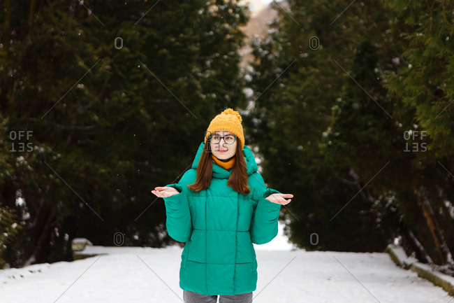 The young female woman in a green winter jacket is walking between the snowy trees in the park or forest that is full of snow