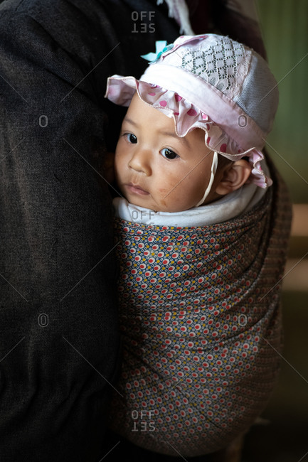 AKHA HILL TRIBE, HOKYIN VILLAGE, MYANMAR - 22 January 2019: Close up portrait of hill tribe baby in traditional wrap sling on mothers back.
