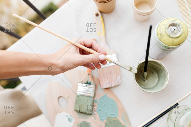 Top view of cropped unrecognizable woman holding brush near various paint and brushes placed on table in creative workshop